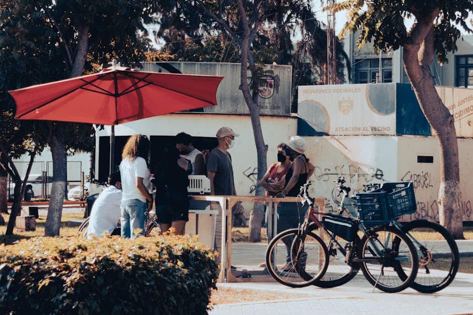 People socializing outdoors under a red umbrella in a park setting with bicycles nearby.