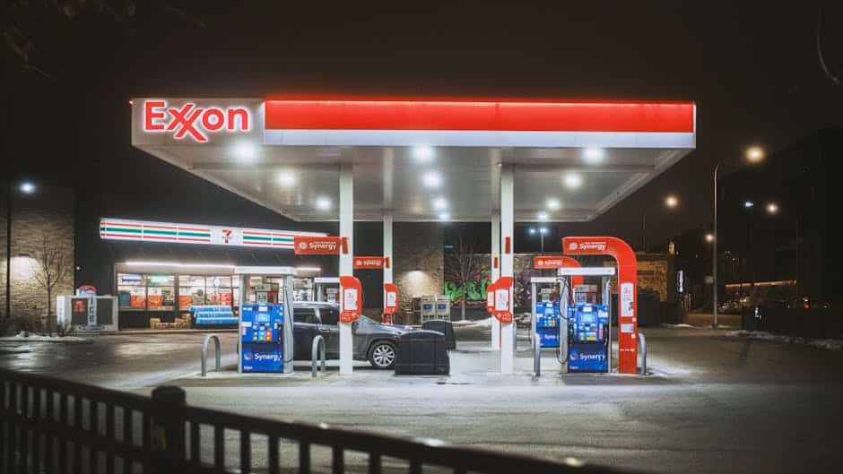 A brightly lit Exxon gas station and 7-Eleven store at night, featuring fuel pumps and a parked car.